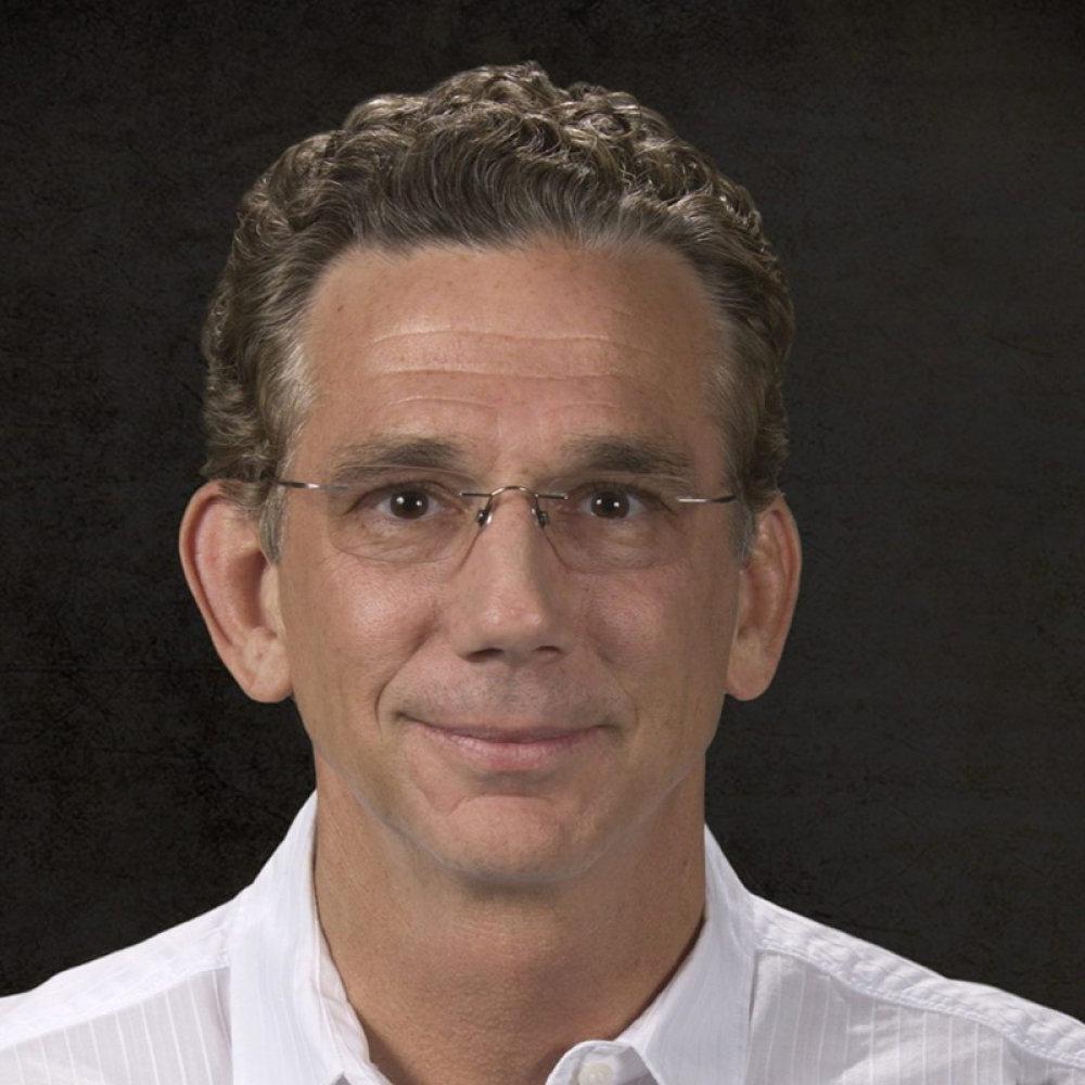 Man with dark curly hair wearing glasses and white button up dress up shirt in front of black background