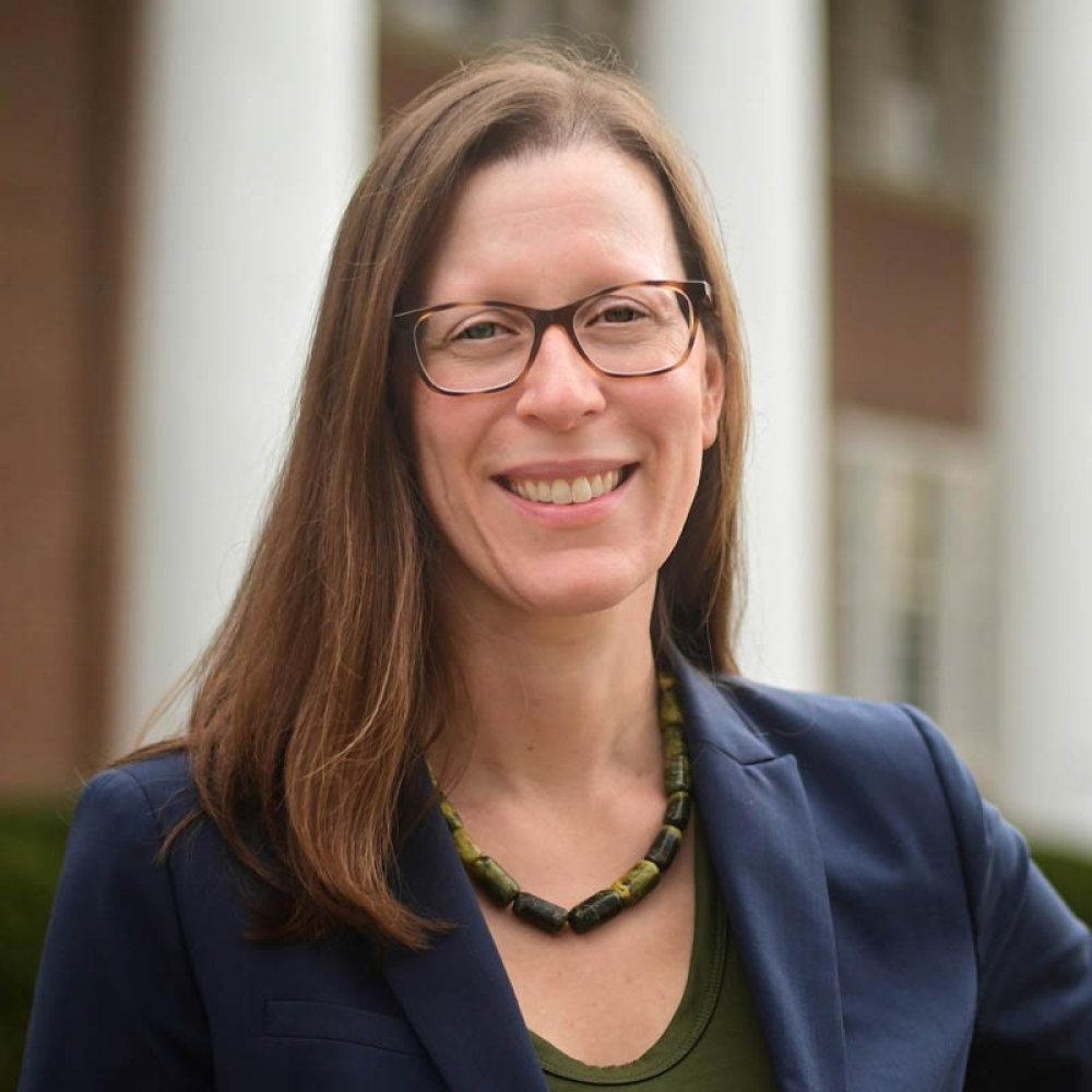 Lady with dark brown shoulder length hair with glasses, wearing navy blazer and dark top in front of Old Centre