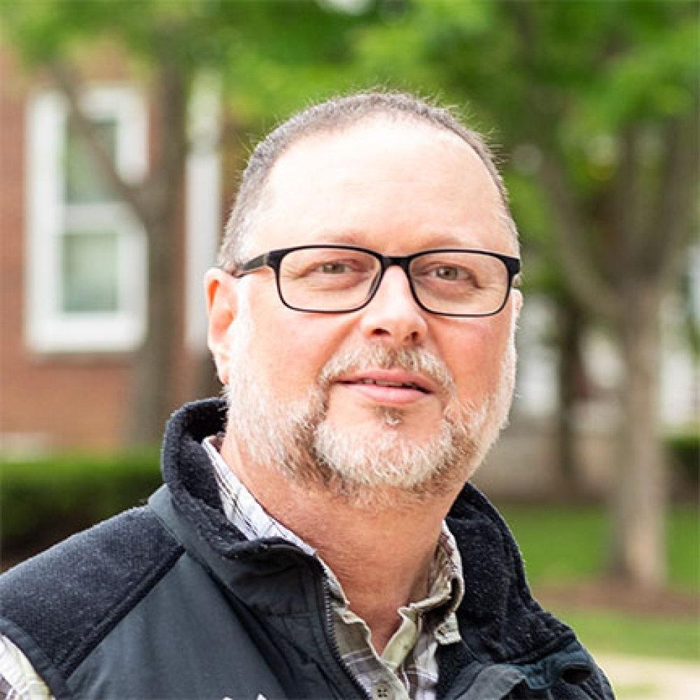 Man with short dark hair with beard wearing glasses, dark vest and plaid dress shirt